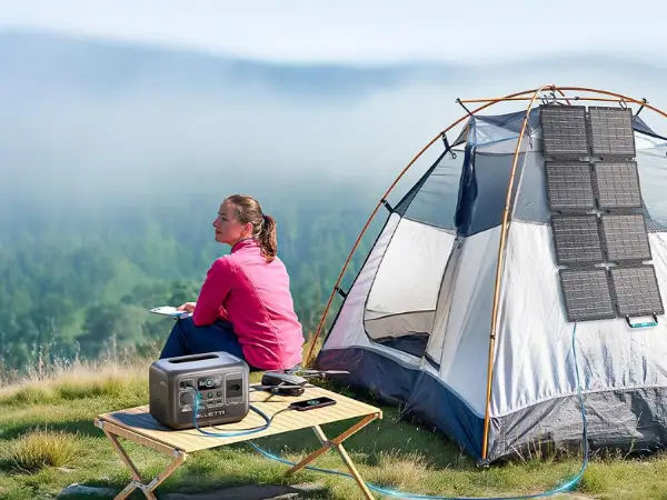 Painel solar portátil dobrável no chão, recarregando uma estação de energia ao lado de uma barraca de camping em um dia ensolarado.