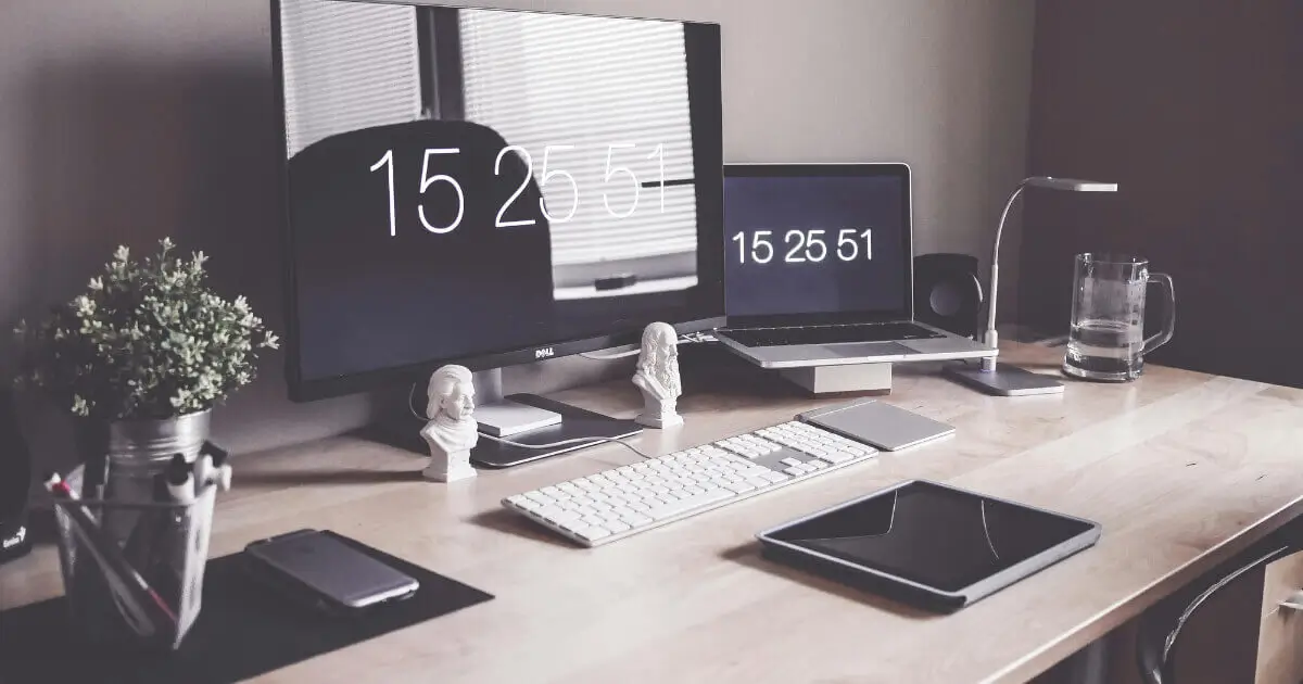 Mesa de um home office moderno e organizado com um monitor grande, laptop, teclado sem fio e outros acessórios.