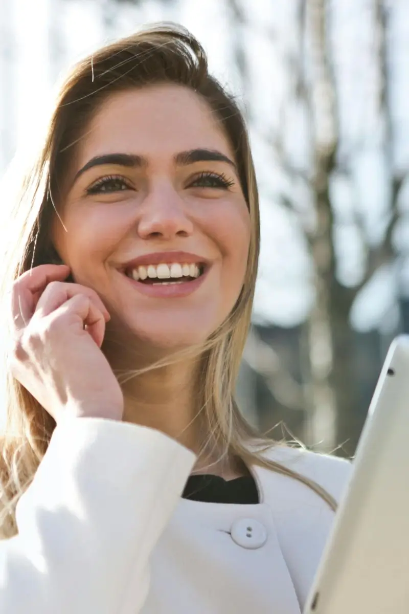 Mulher jovem e sorridente de cabelos loiros usando um tablet em um ambiente externo e ensolarado.